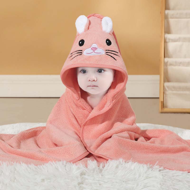 Baby wrapped in a pink towel with an animal hood, sitting on a fluffy white rug against a beige background. The towel hood features a cute embroidered face and ears.