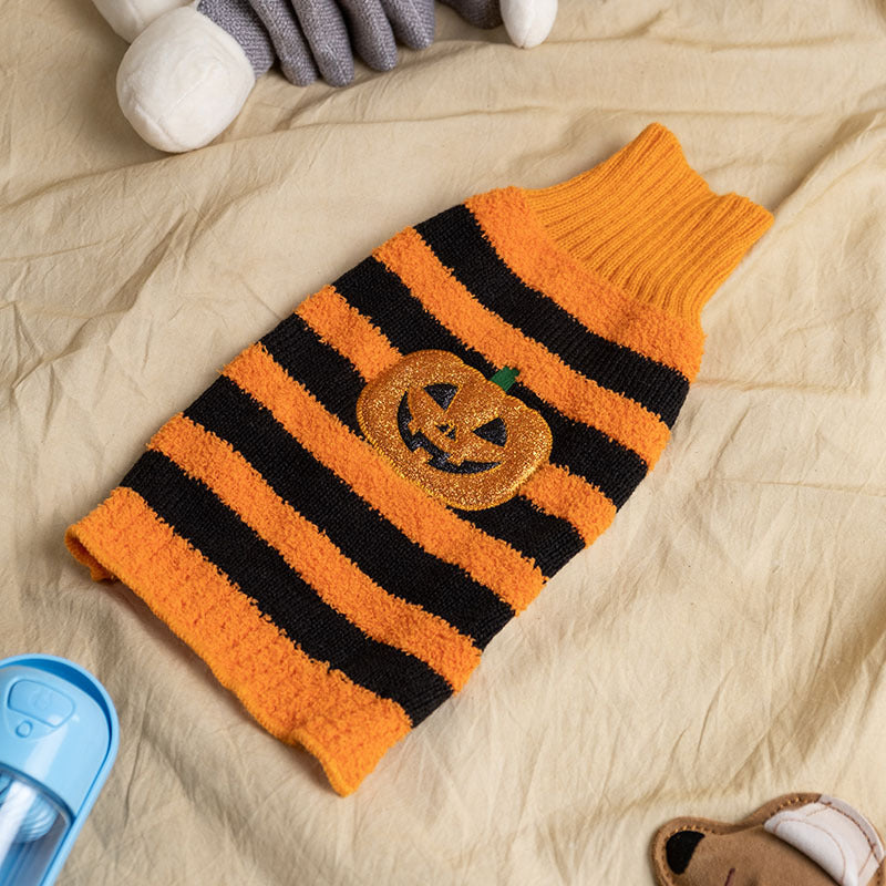 Striped orange and black dog sweater with a glittery pumpkin design on a cream fabric background. A blue plastic item and stuffed toy are partially visible nearby.