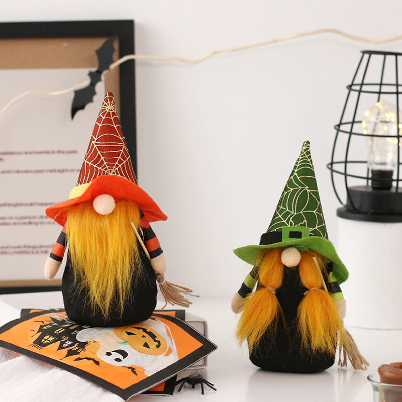 Two Halloween gnomes with long orange beards sit on a table. The left gnome wears an orange spiderweb hat and holds a broom, while the right gnome has a green spiderweb hat. Around them are Halloween-themed cards, a black cage lantern, and bat decoration on the wall.