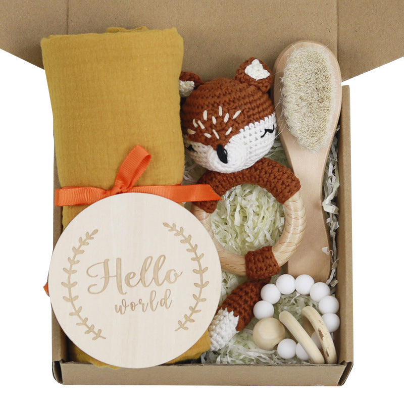 A baby gift box contains a yellow blanket with an orange ribbon, a wooden brush, a crocheted brown animal toy, a white teething ring, and a "Hello World" wooden plaque.