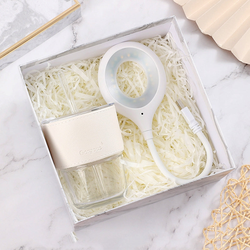 Gift box containing a glass mug with a white sleeve labeled "Gorgeous" and a white USB LED ring light, set against shredded white padding on a marble surface.