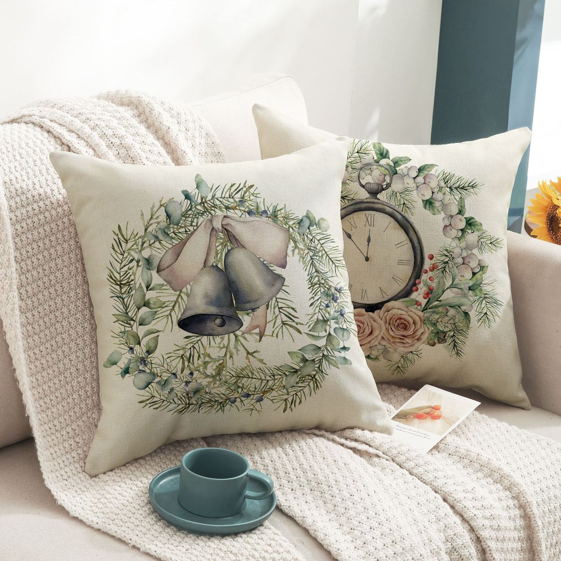 Two decorative pillows on a beige sofa feature festive designs: one with bells and a wreath, the other with a clock and flowers. A teal cup and saucer sit on a knit blanket.