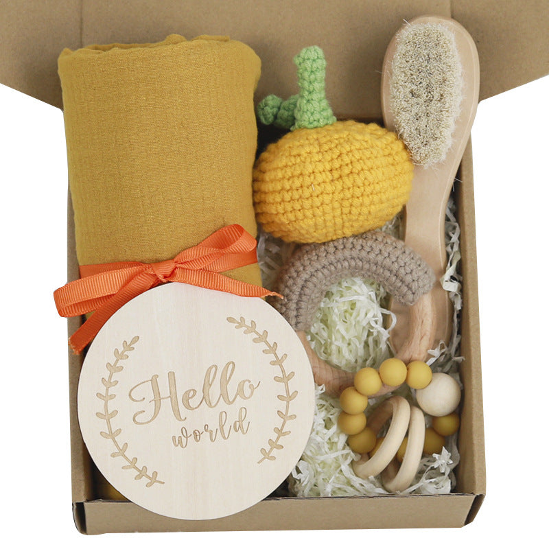 Gift box with a yellow blanket, wooden brush, crocheted pumpkin, rainbow toy, and teething ring on shredded paper. A wooden plaque reads "Hello world" with floral accents.