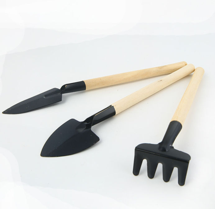 Three miniature gardening tools on a white background: a trowel, a shovel, and a rake, each with light wooden handles and black metal heads.