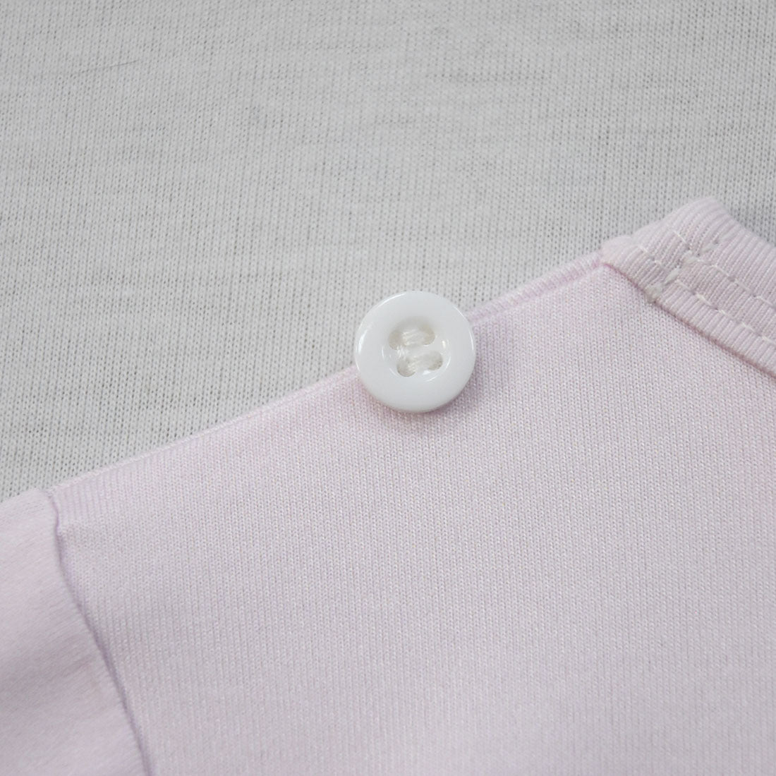Close-up of a white button on a light pink fabric, sewn neatly with visible stitching. The background is a soft, textured gray material.