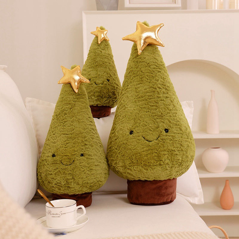 Three plush Christmas trees with smiling faces and gold star tops are placed on a sofa. A teacup sits nearby, and soft decor is visible in the background.