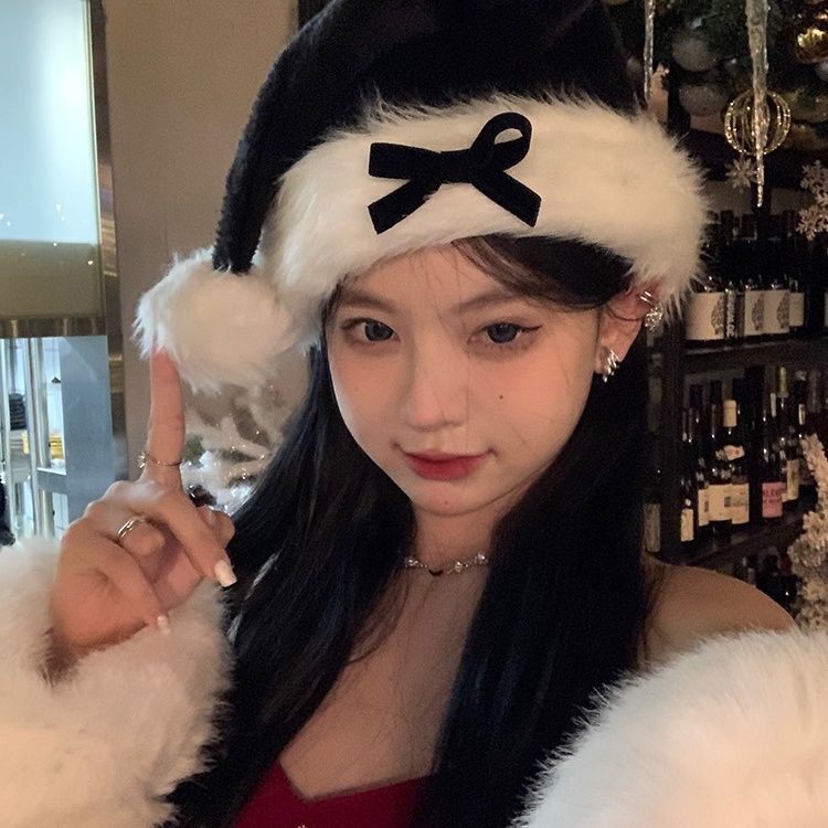 Woman in a Santa hat and red outfit poses with one finger pointing up. She is surrounded by festive decor, including a decorated Christmas tree and wine bottles.