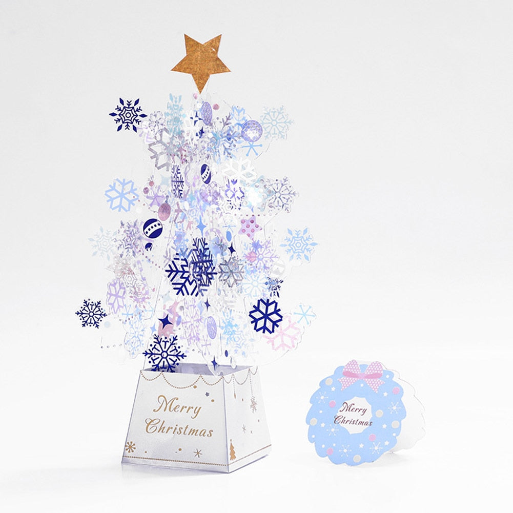 Pop-up Christmas card with abstract snowflake tree topped by a gold star. Base reads "Merry Christmas." Additional small card with "Merry Christmas" and a pink bow lies nearby.