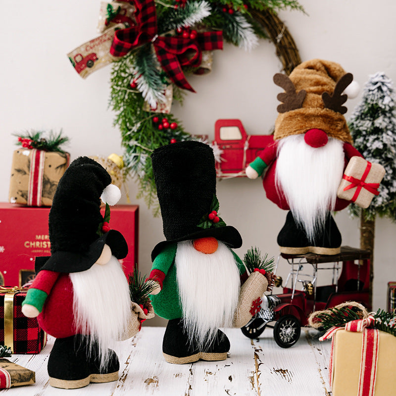 Three festive gnome figurines with long white beards, wearing holiday hats, hold gifts and greenery. They stand among wrapped presents and a decorated wreath on a rustic table.