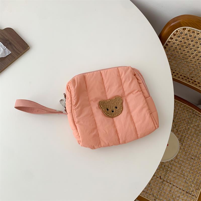 A peach quilted pouch with a bear patch and a wrist strap lies on a round white table next to a rattan chair and a small wooden board with a tissue.