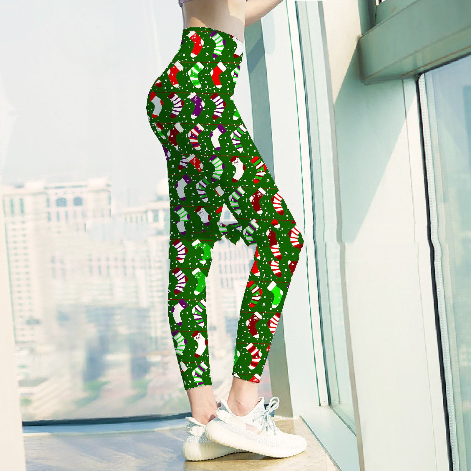 Woman wearing festive green leggings with colorful Christmas stocking pattern, standing near a large window. Sneakers complete the casual look with a cityscape view outside.