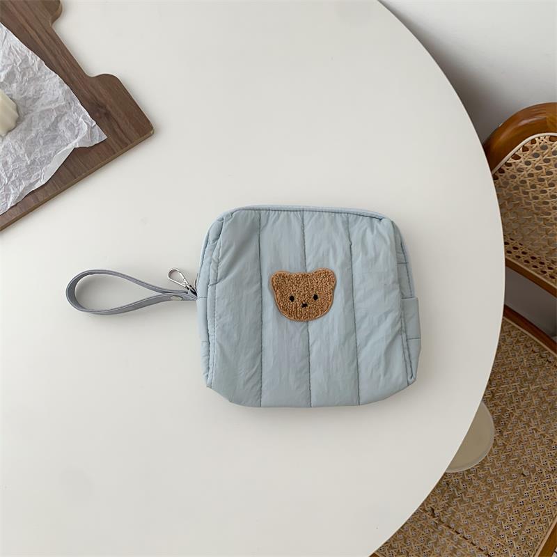 Light blue quilted pouch with a bear patch, resting on a white round table. A woven chair is partially visible in the foreground, with a wooden board with paper nearby.