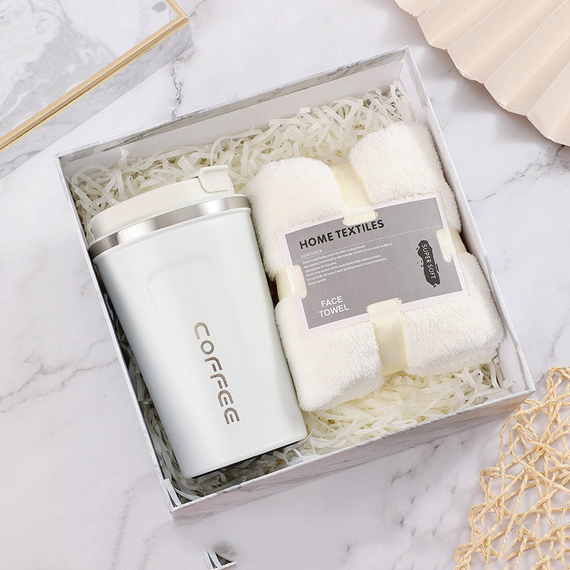 Gift box with a white insulated coffee tumbler and a set of soft, white face towels, nestled in decorative shredded paper, set on a marble surface.