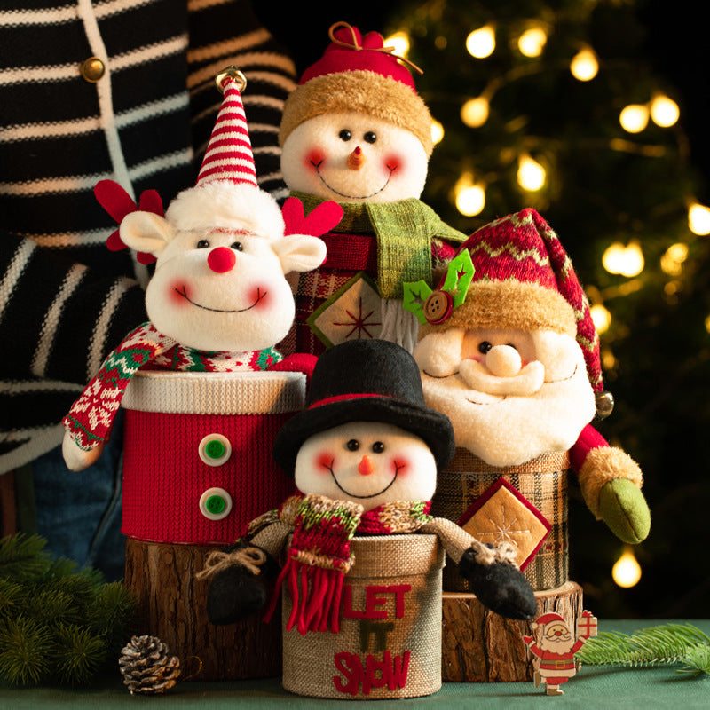 Festive holiday decorations featuring smiling plush snowmen, a reindeer, and Santa in colorful hats and scarves, set against a backdrop of glowing string lights and greenery.