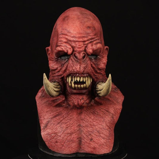 A detailed, red, monstrous bust with a bald head, wrinkled skin, sharp teeth, tusks, and dark eyes against a black background.