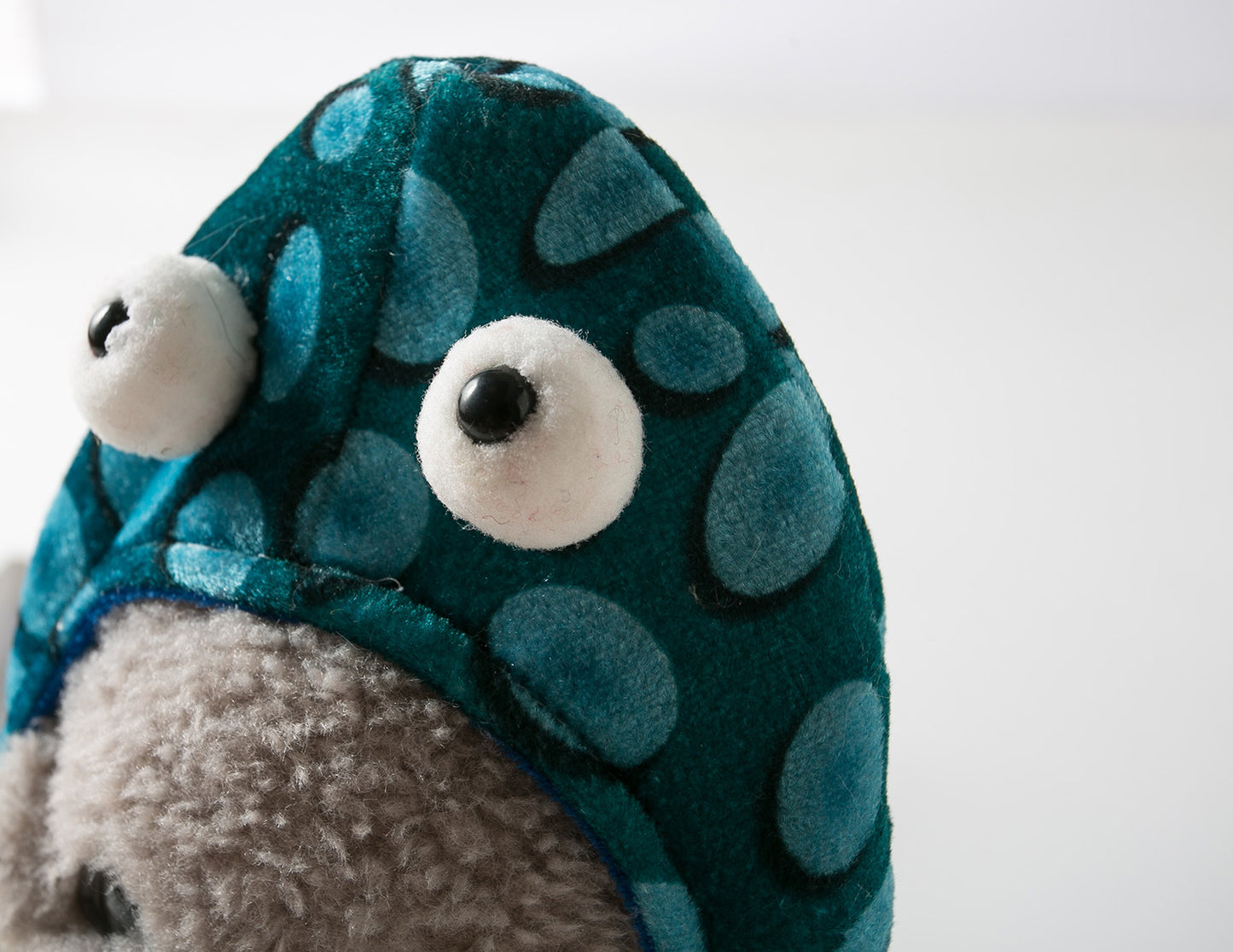 Close-up of a plush toy with big eyes on a blue, polka-dotted hood, over a textured gray face and a white background.