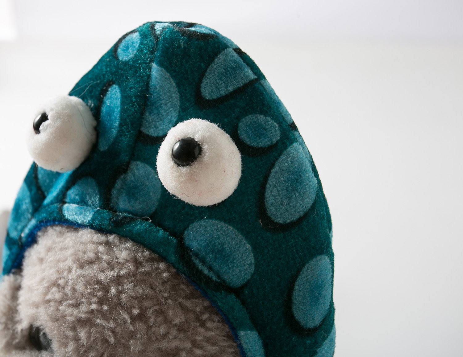 Close-up of a plush toy with big eyes on a blue, polka-dotted hood, over a textured gray face and a white background.