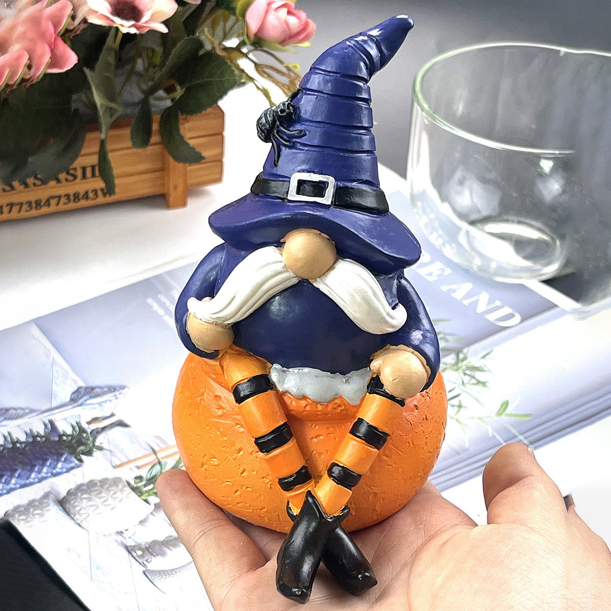 A whimsical gnome figurine wearing a purple witch hat and orange-striped leggings sits on a hand, with a plant and glass cup in the background.