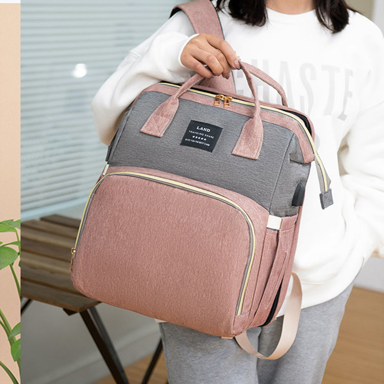 A person wearing a white sweatshirt holds a stylish grey and pink backpack with a front pocket and black label. The setting includes a plant and wooden bench in the background.