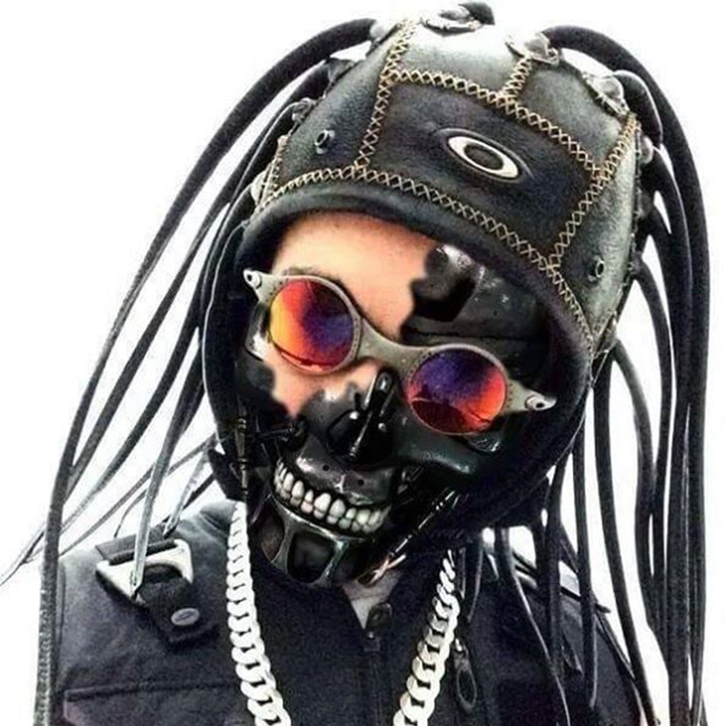 Person wearing a black mask resembling a skull, with reflective sunglasses, a leather helmet adorned with tubes, and a thick chain necklace against a plain background.