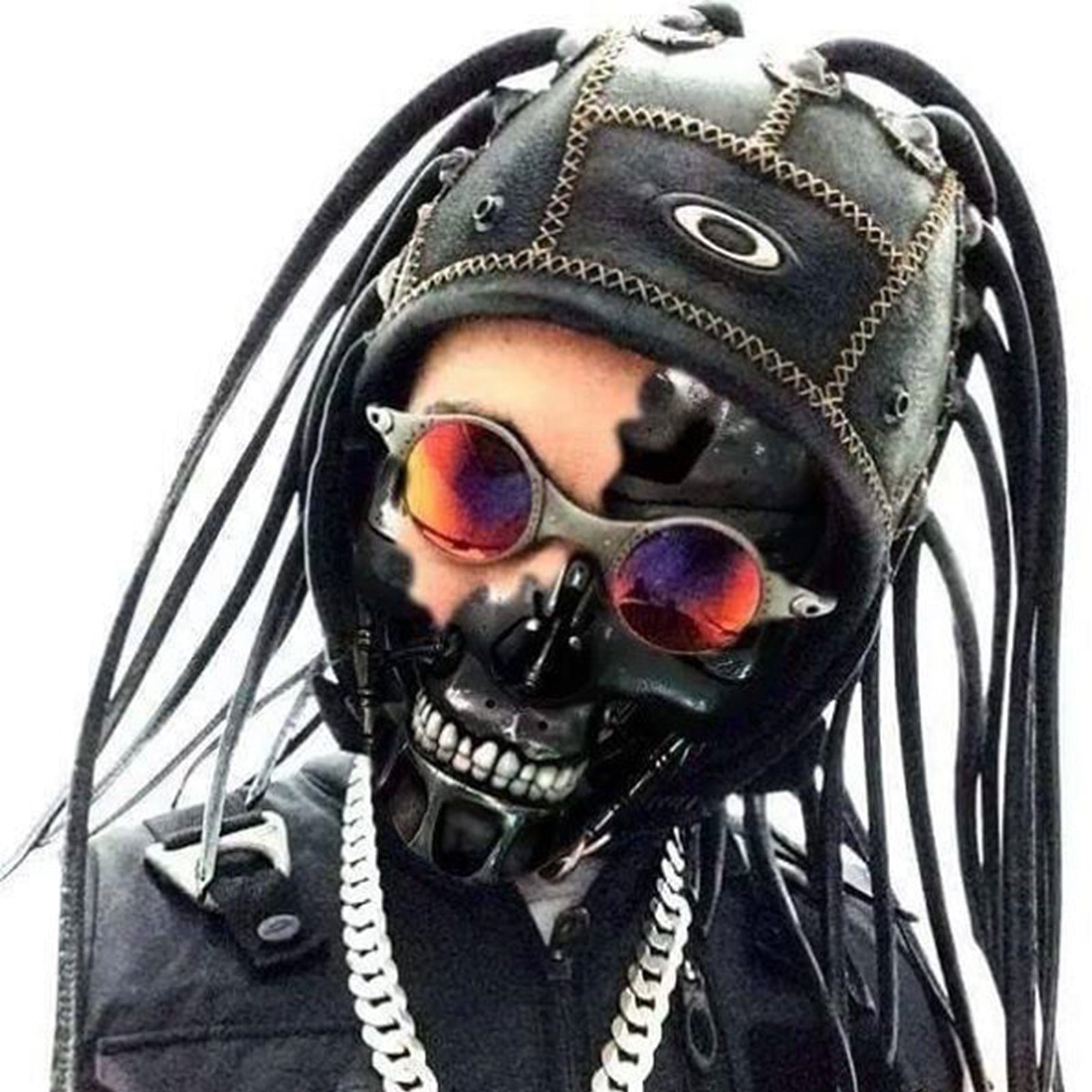 Person wearing a black mask resembling a skull, with reflective sunglasses, a leather helmet adorned with tubes, and a thick chain necklace against a plain background.