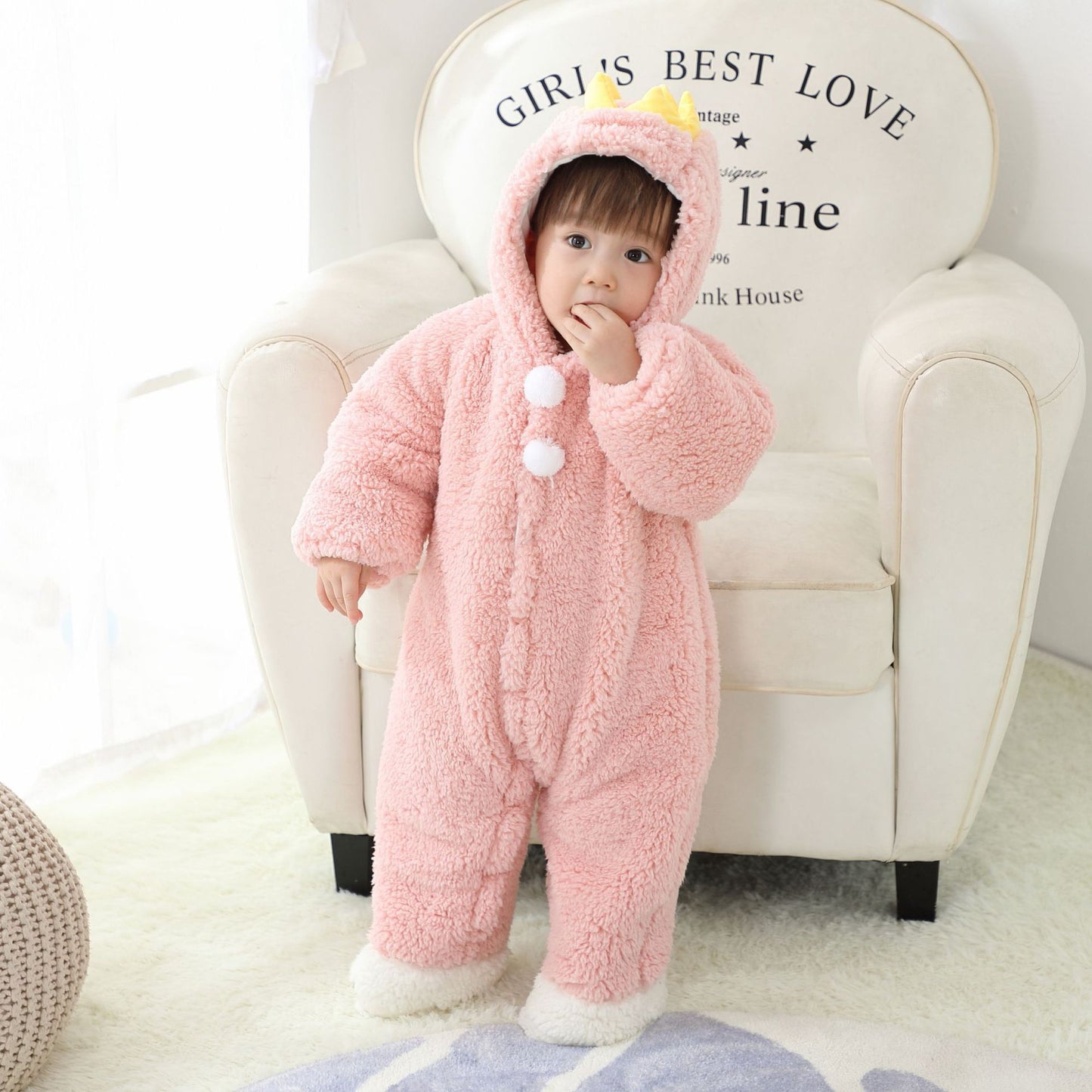 A toddler in a fluffy pink onesie with a hood stands in front of a white armchair. The onesie has white pom-poms and yellow crown-like decorations on the hood.