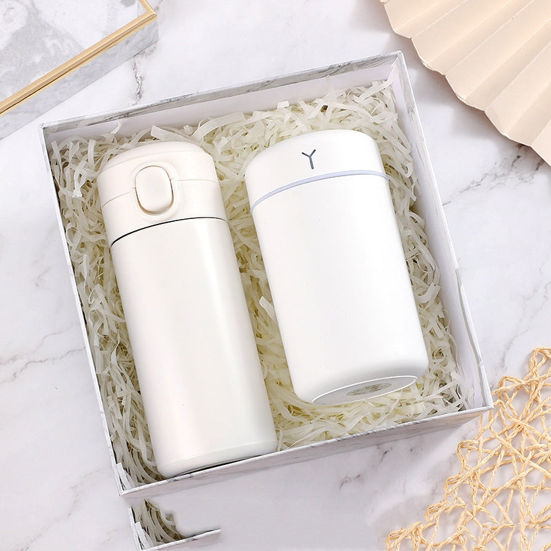 White insulated tumbler and cylindrical container in a box with shredded paper on a marble surface, alongside decorative elements.