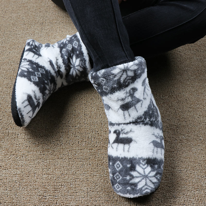 Person wearing cozy, fluffy slippers with black and white reindeer and snowflake patterns, sitting cross-legged on a beige carpet.