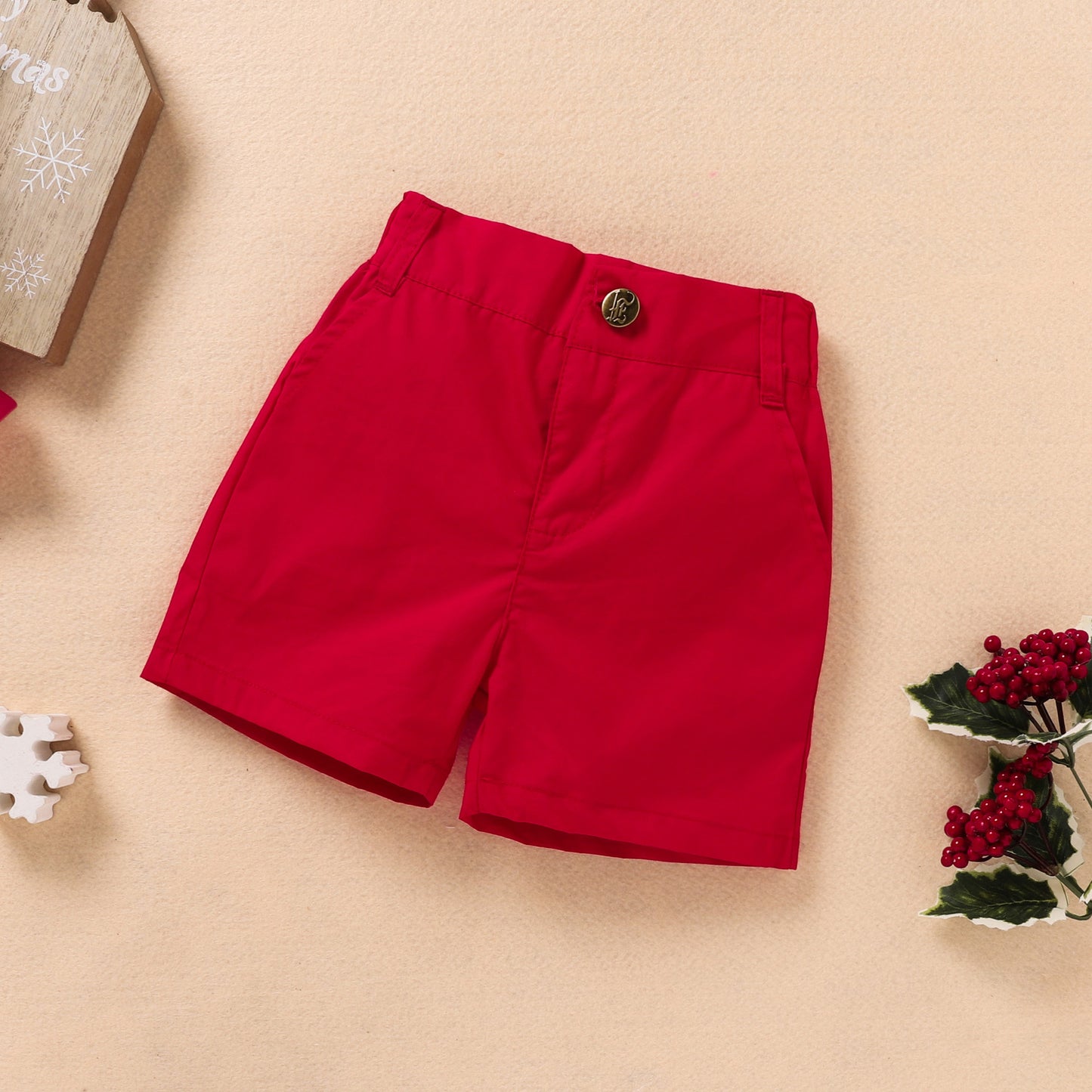 Bright red shorts with a brass button, set against a beige background. Decorative holly and snowflakes surround the shorts, suggesting a festive holiday theme.