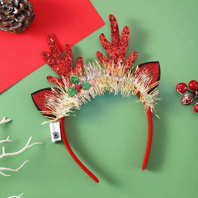 Red glitter reindeer antler headband with tinsel decoration, holly leaves, and berries on a green surface. Nearby are a snowy pine cone, white branch, and red berries.