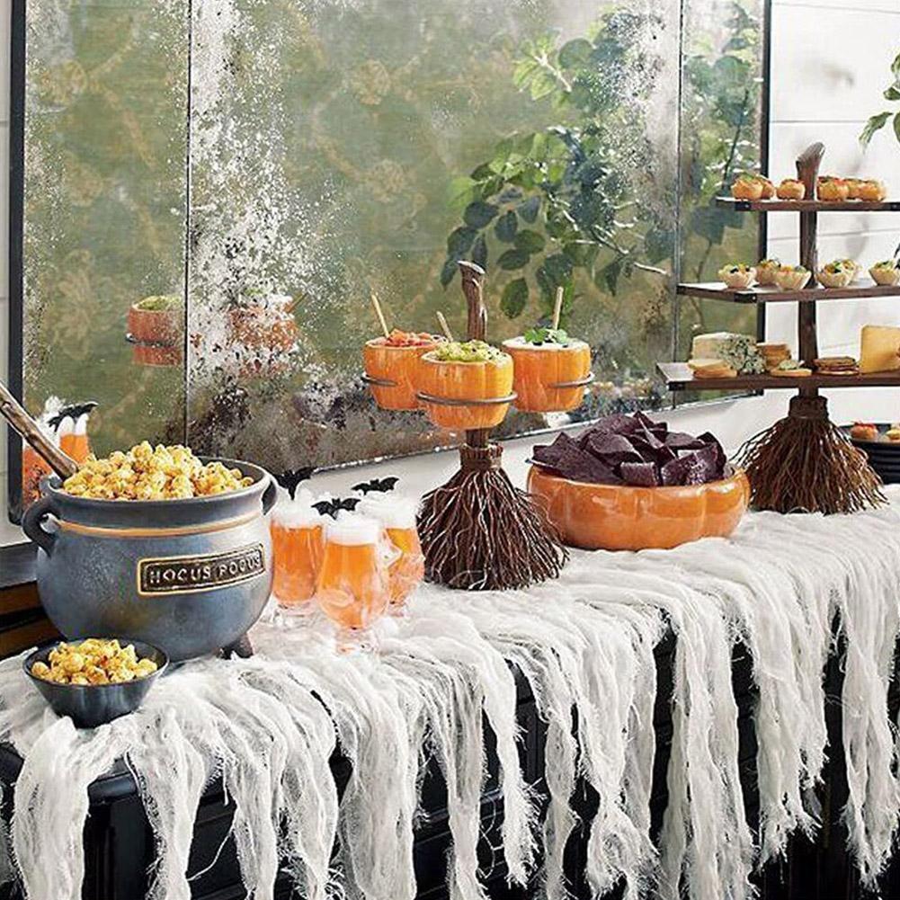 Halloween-themed table with a cauldron of popcorn, orange drinks with bat straws, pumpkin bowls of snacks, purple chips, and layered pumpkin trays. Draped with white cloth.