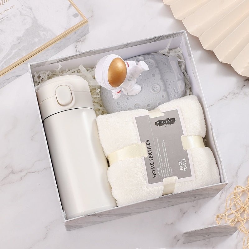 A gift box containing a white insulated tumbler, a rolled white towel labeled "Home Textiles," and a small astronaut figurine on a grey moon surface, all set on a marble background.