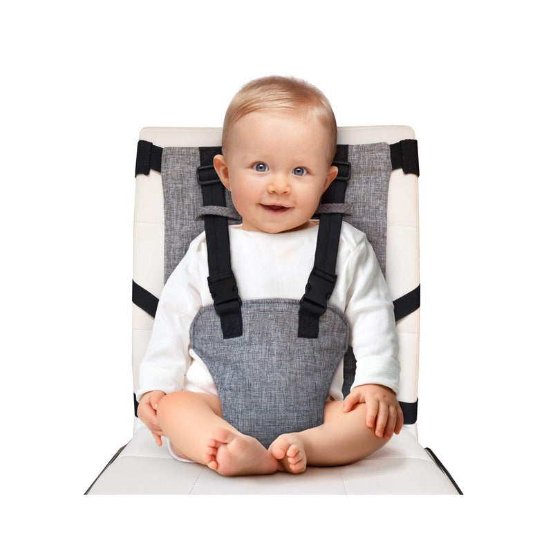 A smiling baby in a white shirt is seated on a gray booster seat with black straps, looking directly at the camera. The seat is placed against a white background.