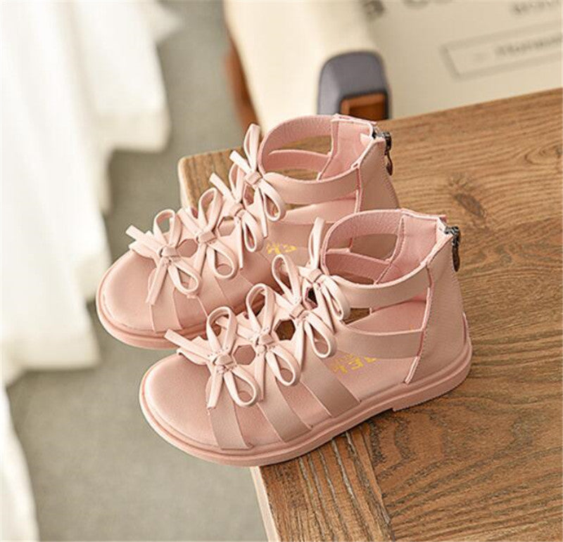 Pink gladiator sandals for toddlers with multiple straps and decorative bows on top, featuring back zippers. Sandals are placed on a wooden surface, viewed from above.