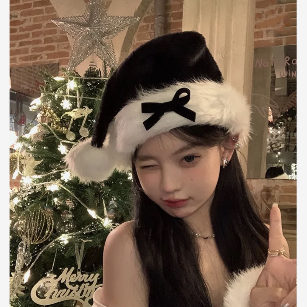 Young woman in a black Santa hat with a bow, winking and pointing upwards. A Christmas tree with lights and a star topper is in the background.