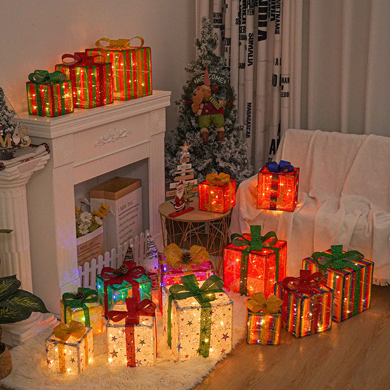 A cozy Christmas setting with illuminated, colorful gift boxes under a decorated fireplace and next to a small Christmas tree and sofa, creating a festive atmosphere.