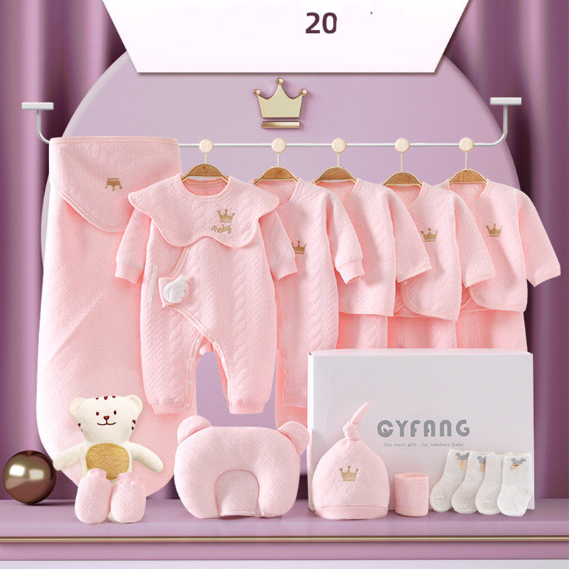 A baby gift set featuring pink clothing with crown embroidery, including a onesie, jackets, and socks. Also shown are a bear plush, pillow, hat, and a branded box.