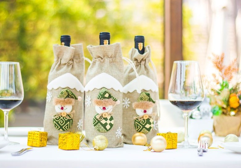 Three wine bottles in burlap holiday bags with snowman designs are on a table with red wine glasses, gold ornaments, and a festive centerpiece.