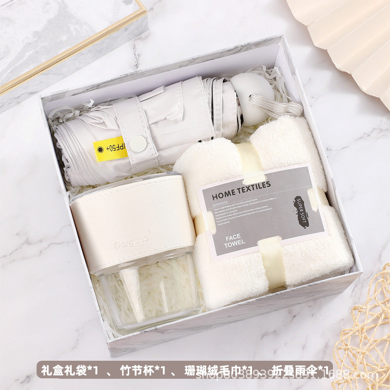 Gift box containing a folded umbrella, soft face towel, and a white tumbler cup with lid. All items are neatly arranged against a marble background.
