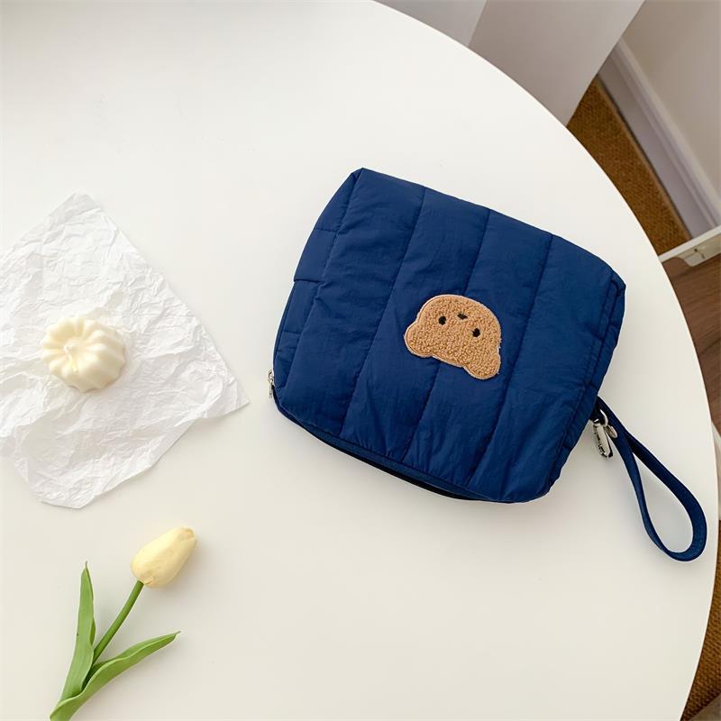A blue quilted bag with a bear patch and wrist strap on a white table. Nearby are a crumpled paper holding a white pastry and a single pale yellow tulip with green leaves.