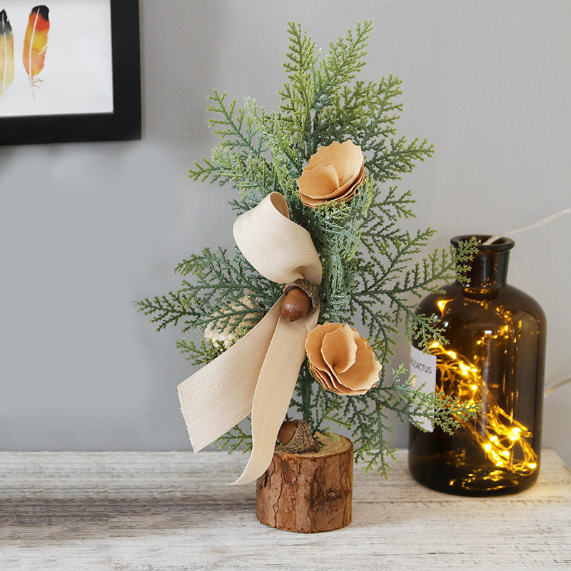 Miniature evergreen tree with beige paper roses, a ribbon, and an acorn, set on a wood slice. Nearby is a dark glass bottle filled with string lights. Art on the wall.