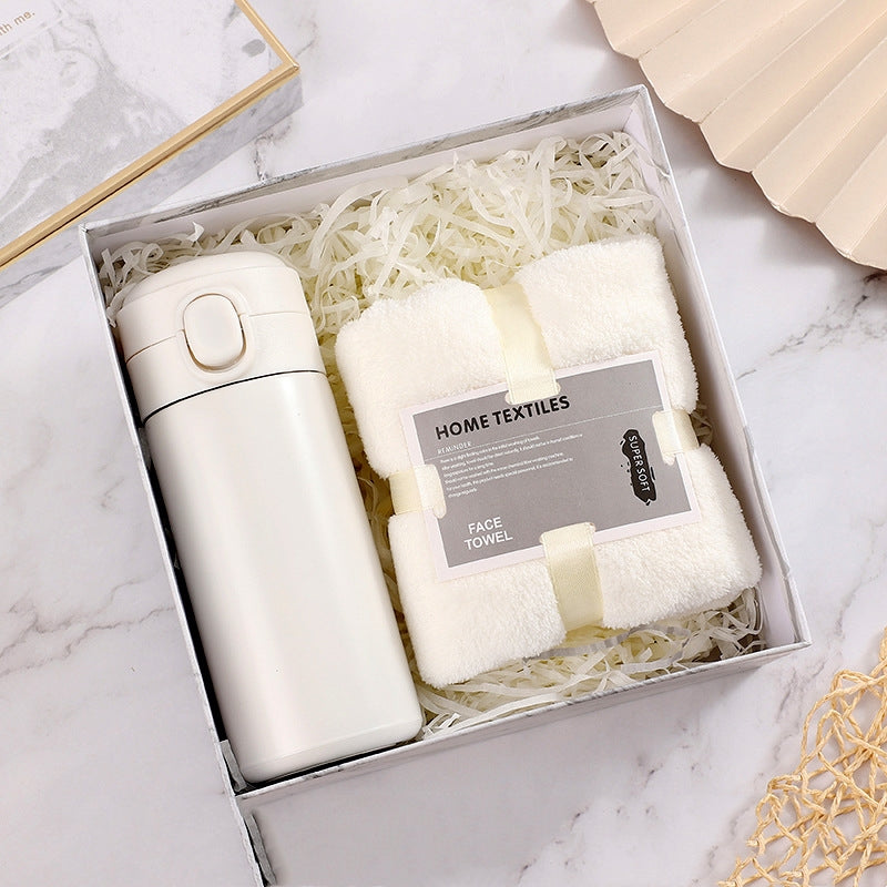 A white gift box contains a sleek insulated water bottle and a neatly folded white face towel labeled "Home Textiles," set on shredded paper filler on a marble surface.