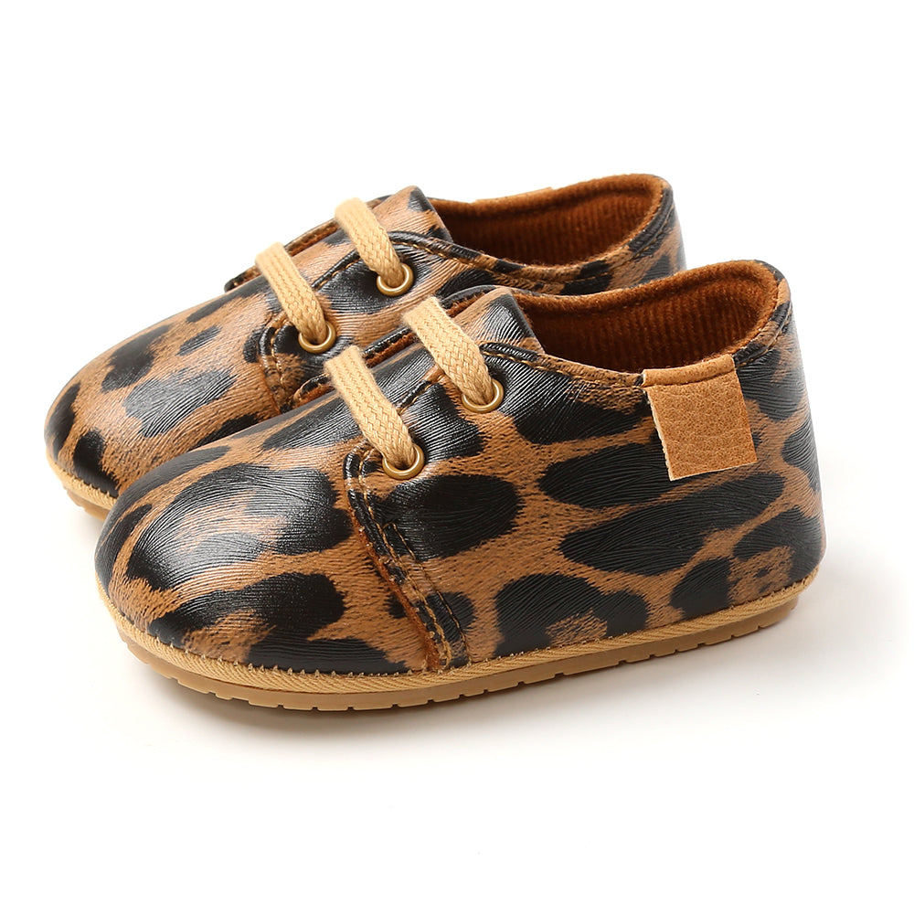 Brown baby shoes with a black leopard print pattern, featuring tan laces and soft soles. The shoes are styled to look like miniature sneakers and are positioned side by side.