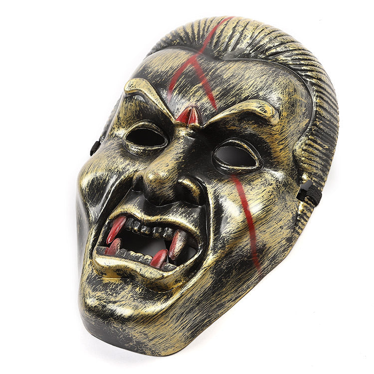 A fierce metallic mask with exaggerated features, red scars, and prominent fangs, resembling a vampire. The mask has a distressed gold and black finish.