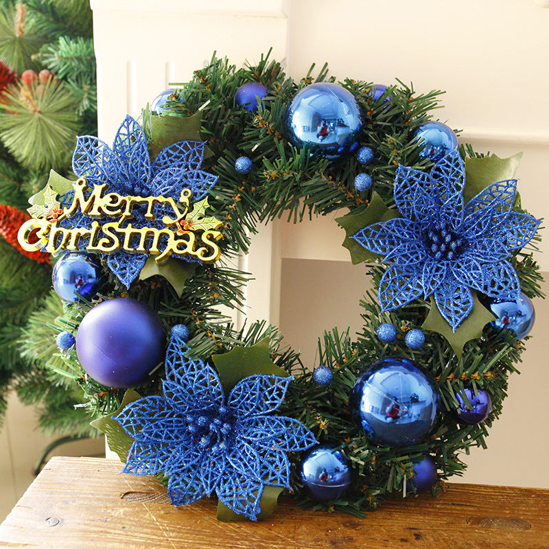 Christmas wreath with blue ornaments, poinsettia flowers, and a "Merry Christmas" sign, displayed on a wooden surface with a hint of green foliage in the background.