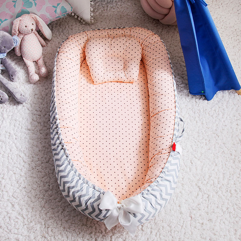 A cozy baby lounger with peach polka-dot fabric and gray chevron sides, featuring a small pillow and white bow. It’s set on a plush rug with plush toys and a blue canopy nearby.
