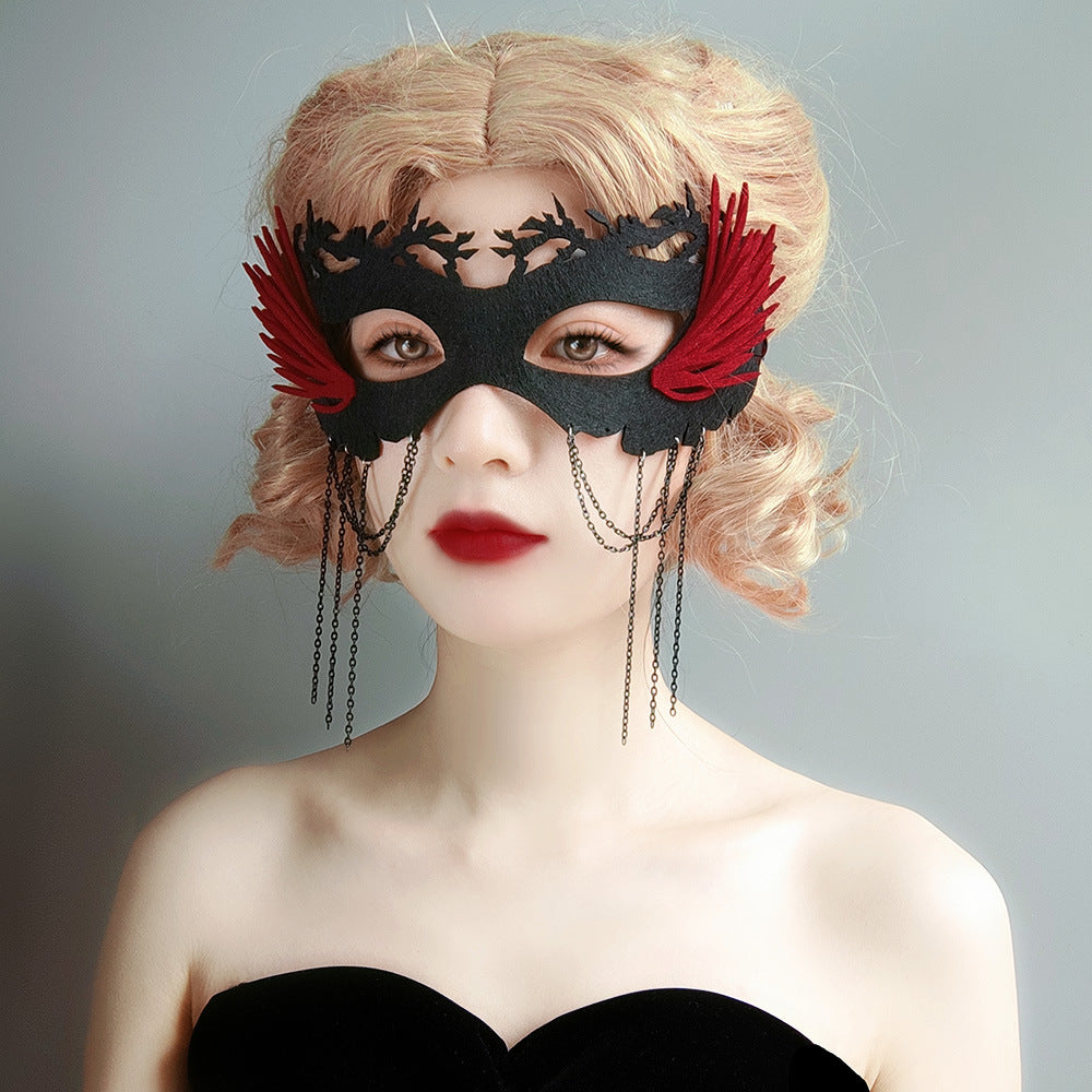 Woman with curly blonde hair wears an ornate black mask adorned with red feathers and dangling chains, complementing her strapless black top against a soft gray background.