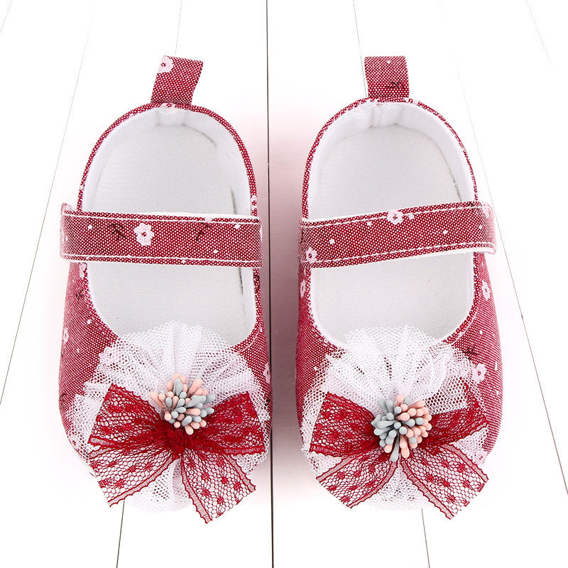 Red baby shoes with white polka dots and floral patterns, featuring white lace bows with pink and blue decorative beads on toes, set against a white wooden background.