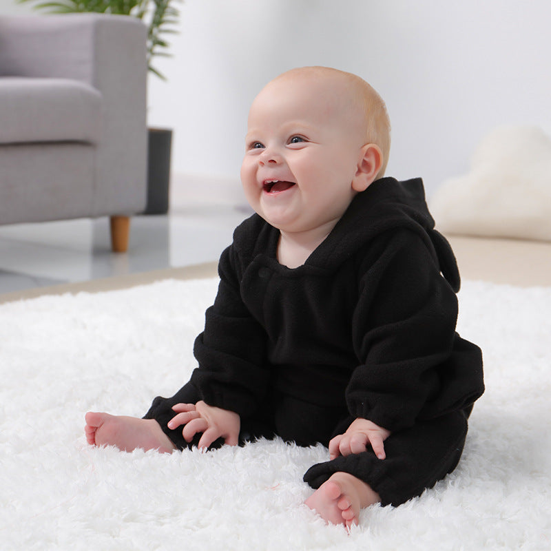 A smiling baby in a black onesie sits on a fluffy white rug in a bright living room. A gray couch and plant are in the background, creating a cozy atmosphere.