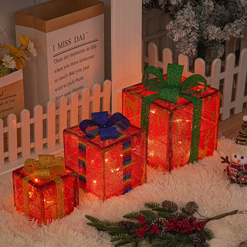Three illuminated red gift boxes with colorful bows sit on a fluffy rug. Nearby are a fence, festive decor, a small snowman figure, and a branch with berries.